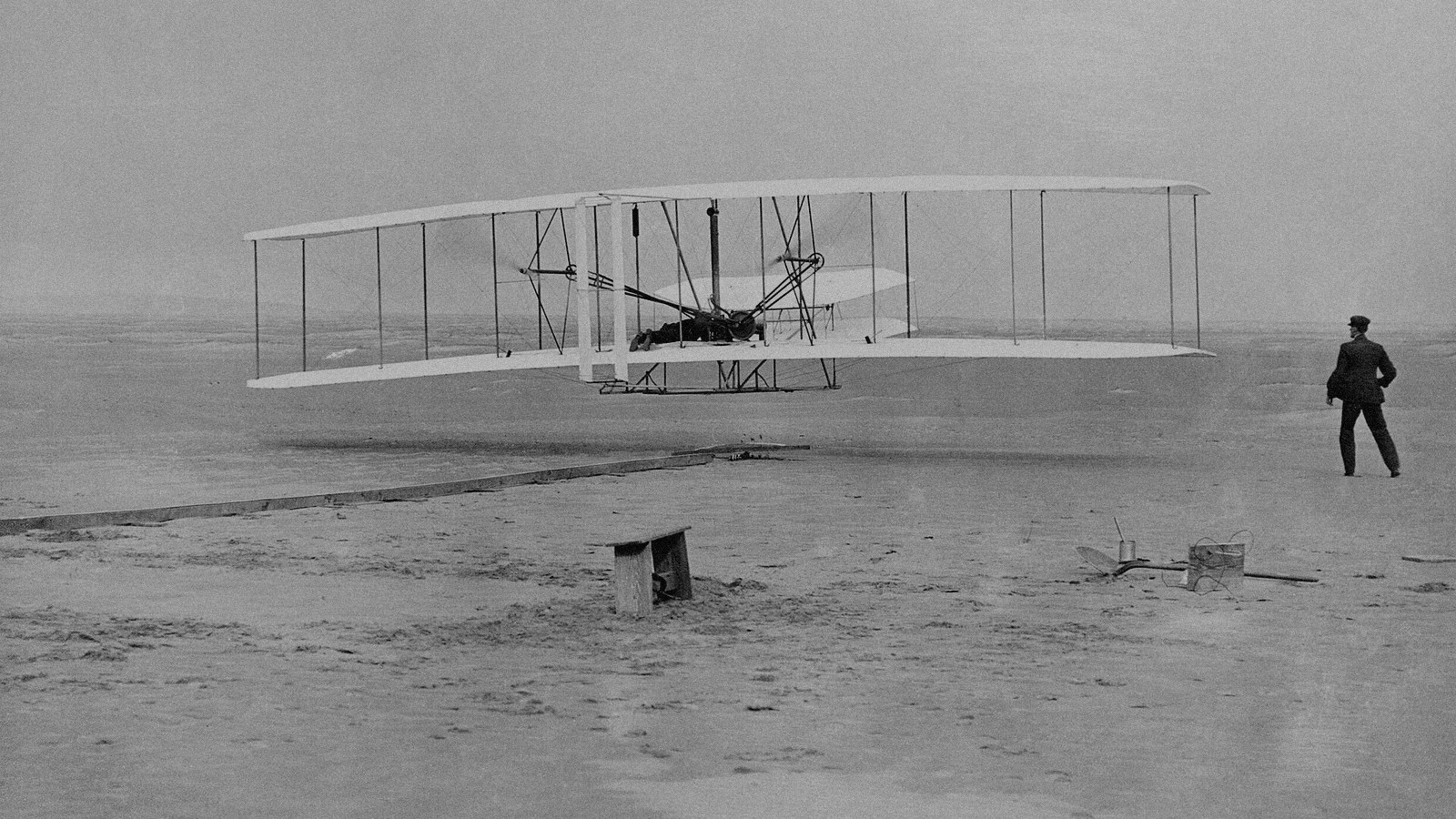 The Wright Flyer lifts off the sand at Kitty Hawk in December 1903 as one brother watches from the right.