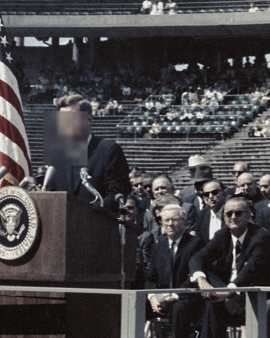 A head of state addresses a stadium crowd from a podium beneath a presidential seal, the speaker's face redacted.